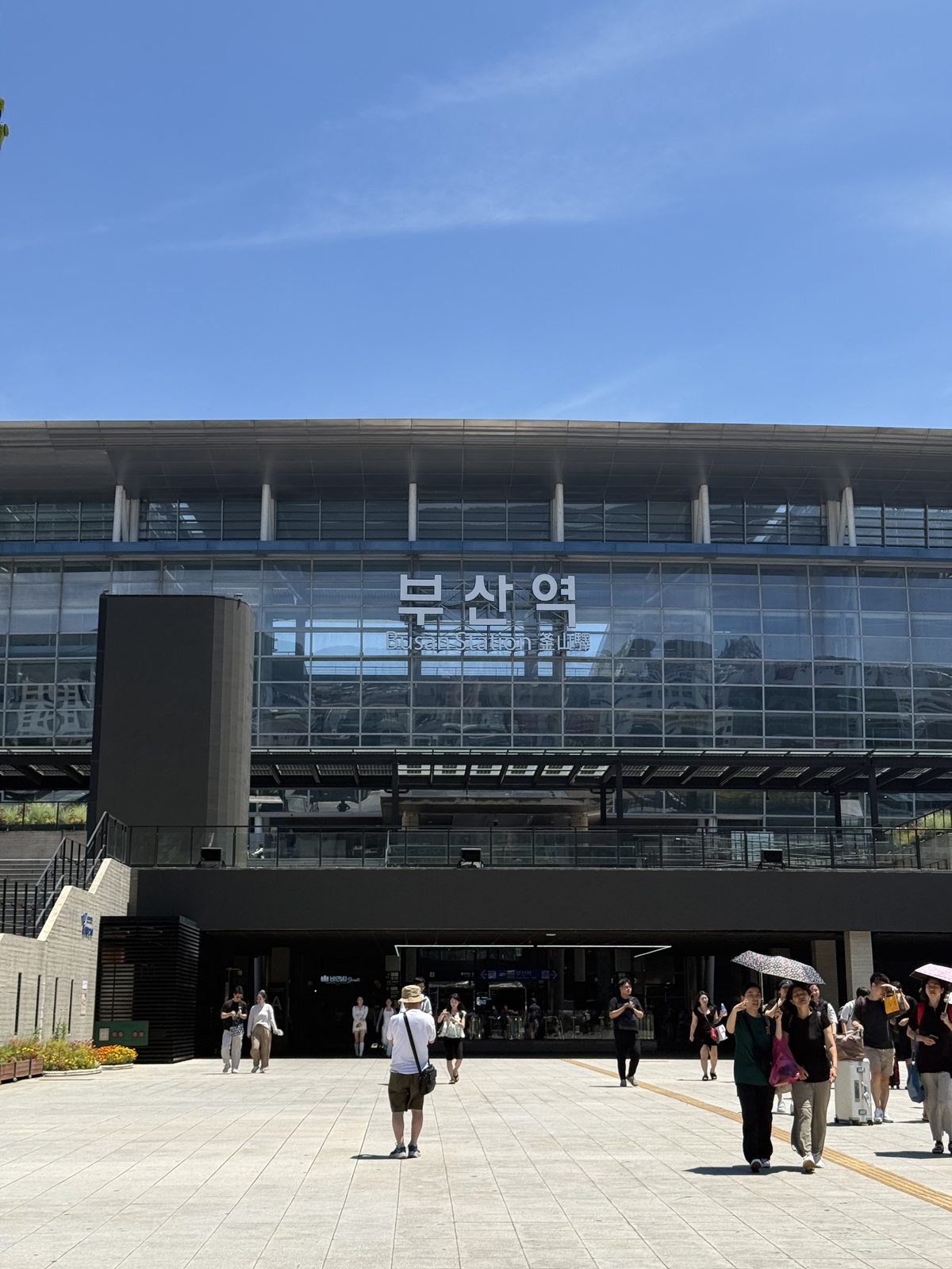 The glass front of Busan Station on a clear day, with travellers crossing the plaza outside