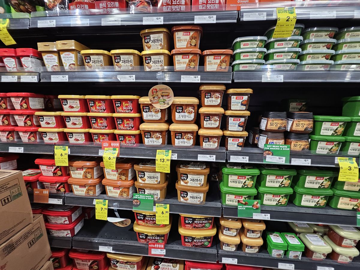 Supermarket shelves stacked with tubs of Korean doenjang (fermented soybean paste) in yellow, brown and red containers, including clam-anchovy doenjang varieties.