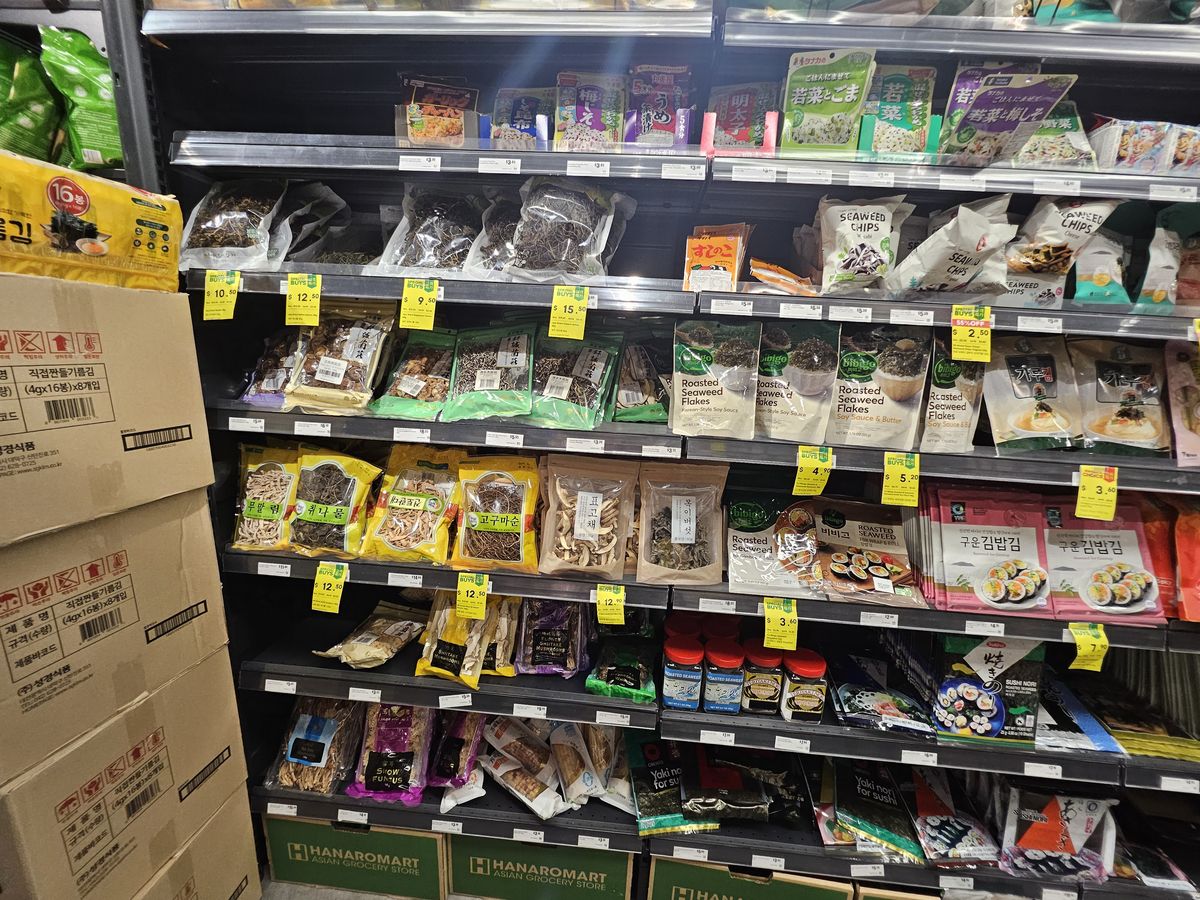 A store shelf of dried Korean vegetables — fernbrake, sweet potato stems, taro stems and mushrooms in clear packs — with seaweed snacks nearby.