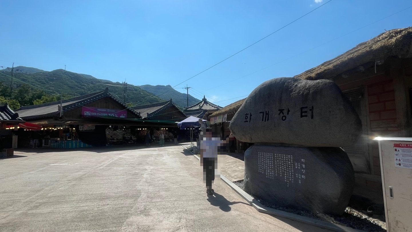 The entrance to Hwagaejangteo market in Hadong, with the engraved stone marker beside thatched-roof and tiled-roof stalls under a clear sky