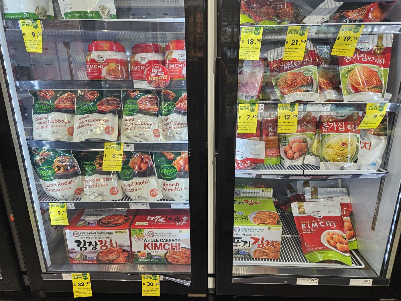 A refrigerated kimchi section in an Australian supermarket: two glass-door fridges stocked with dozens of packaged kimchi varieties — whole cabbage, sliced, radish and white kimchi — with price tags.