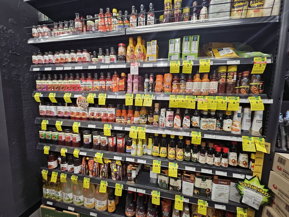 A wall of Korean sauces and condiments in an Asian grocery store: soy sauces, vinegars, syrups, cooking wines and pastes across five shelves with price tags.