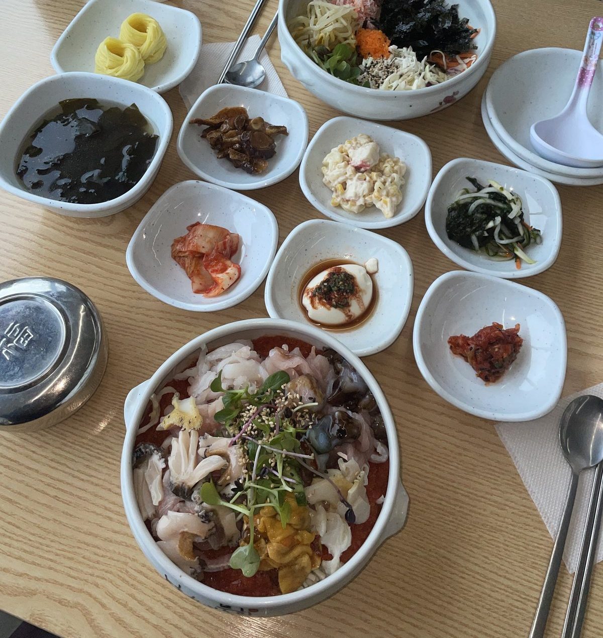 A Korean seafood set meal: a large bowl of assorted raw seafood over rice, a crab bibimbap bowl, and about ten small side dishes including kimchi, seaweed soup, salted seafood and seasoned vegetables on a light wooden table.