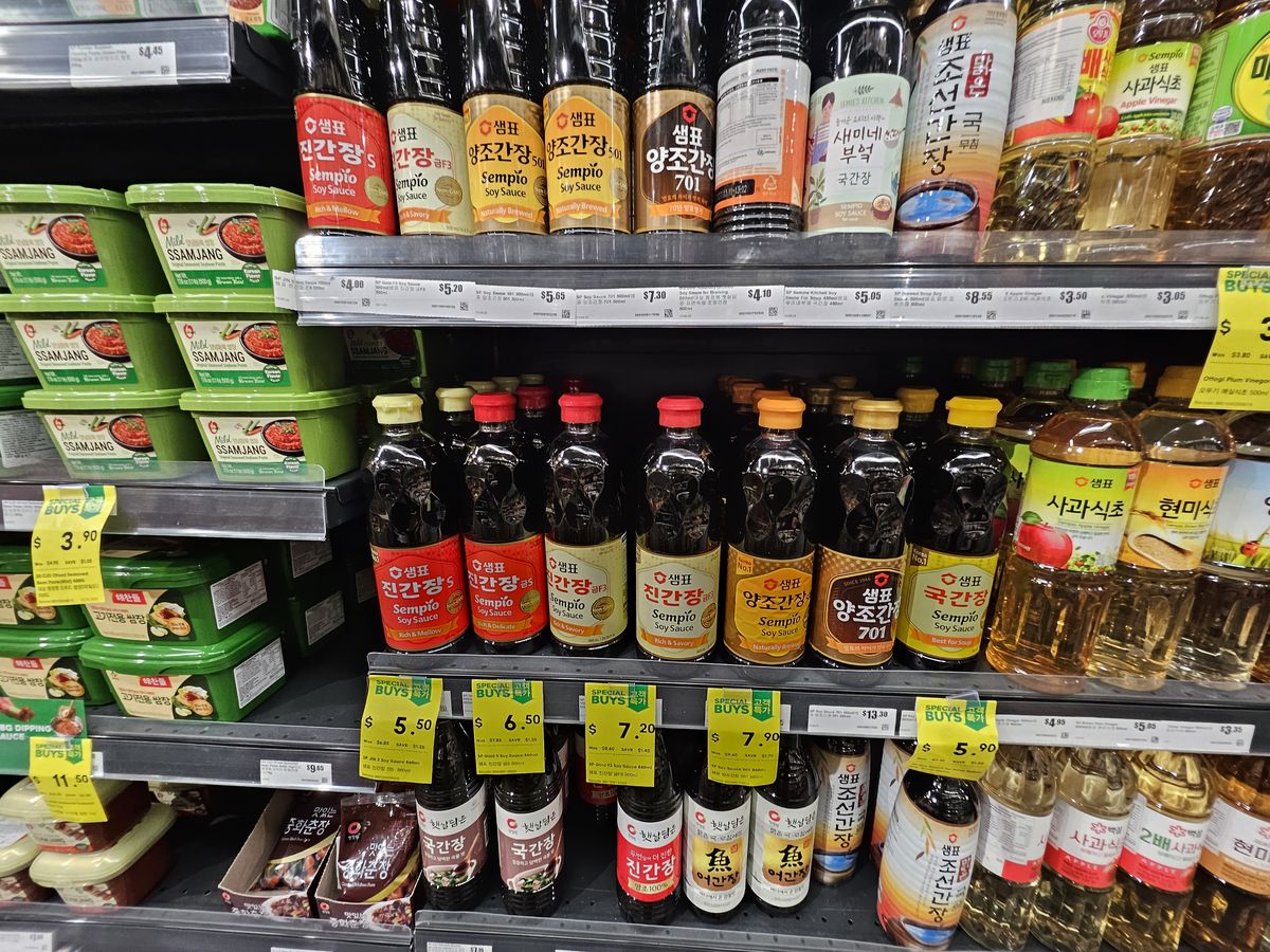 A supermarket shelf of Korean soy sauces — jin ganjang, yangjo ganjang, guk ganjang bottles — beside tubs of ssamjang.