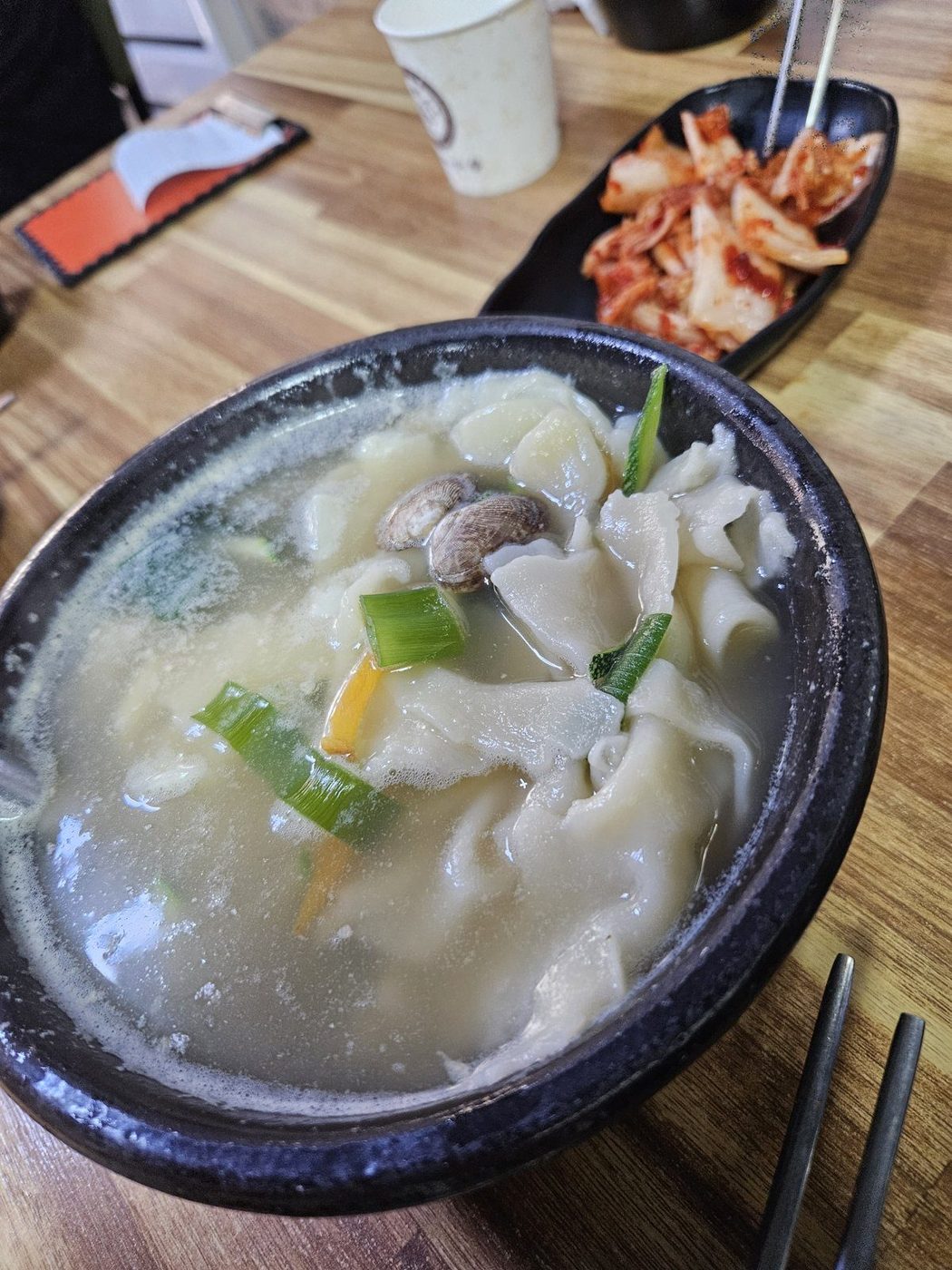 A stone bowl of sujebi — hand-torn wheat dough soup with clams and zucchini in a pale broth — with a side dish of kimchi behind it on a wooden table.