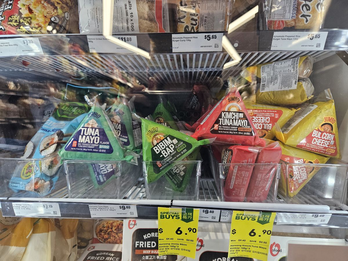 A refrigerated case of Korean triangle kimbap (samgak gimbap) in flavors like tuna mayo, bibimbap and kimchi tuna, with cup rice bowls above.